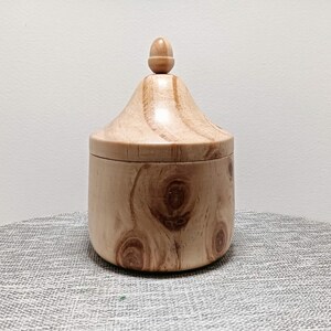 May include: A wooden box with a lid shaped like a rounded acorn. The box is made of light brown wood with a natural grain pattern.