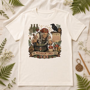 Op de afbeelding: Een gebroken wit t-shirt met een grafische illustratie van een ouder persoon die in een pan roert, een raaf, een kalender en paddenstoelen. De tekst "THE MUSHROOM TIMER LIED" staat op een banner onderaan het kleurrijke, gedetailleerde ontwerp.