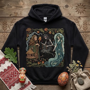 May include: Black hooded sweatshirt with a vibrant illustration of three figures within a decorative frame. The artwork depicts a woman in a long blue dress, a figure holding a torch, and another figure with a small house. Additional decorative elements are present.