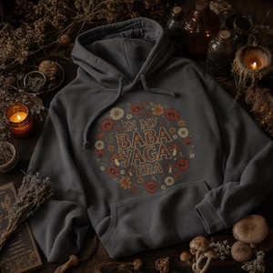May include: A charcoal gray hoodie with a front pocket and drawstring hood. The design features the text "IN MY BABA YAGA ERA" surrounded by a floral wreath with mushrooms, in shades of orange, red, and cream.