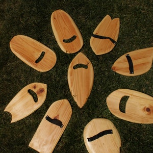 May include: Eight wooden surfboard-shaped objects with black leather handles. The objects are arranged in a circle on a green grassy surface.