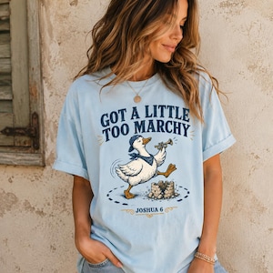 May include: A light blue t-shirt featuring a graphic of a white duck wearing a blue cap and bandana, playing a trumpet while marching near a crumbling stone wall. The text reads "GOT A LITTLE TOO MARCHY" above the duck and "JOSHUA 6" below it.
