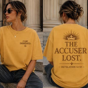 May include: Mustard yellow t-shirt with the text "CASE DISMISSED." and a scale graphic on the front. The back of the shirt features the text "THE ACCUSER LOST." with a crown graphic and the Bible verse Revelation 12:10.