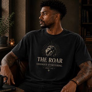 May include: Black t-shirt with a lion graphic and the text "THE ROAR CHANGED EVERYTHING." The design includes a lion's head surrounded by sun rays. The shirt also features the text "AMOS 3:8" and a cross symbol.