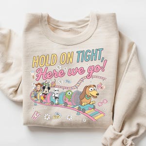 May include: A beige crewneck sweatshirt featuring a colorful graphic of various cartoon characters riding a roller coaster. The text reads, "HOLD ON TIGHT, Here we go!" The design includes small illustrations of flowers, popcorn, and glasses surrounding the coaster track.