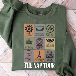 May include: Green sweatshirt with a graphic featuring icons and text from various attractions. The text includes "Carousel of Progress," "Tomorrowland Peoplemover," "Living with the Land," "Hall of Presidents," "Spaceship Earth," "American Adventure," "One Man's Dream," "Impressions de France," "Navi River Journey," and "The Nap Tour."