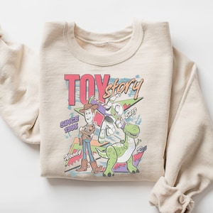 K&ouml;nnte beinhalten: Beiges Sweatshirt mit Rundhalsausschnitt und farbenfroher Grafik von Toy Story-Charakteren. Das Design zeigt Woody, Buzz Lightyear, Rex und den Text "TOY STORY" und "SINCE 1995". Das Sweatshirt ist gefaltet, die &Auml;rmel sind hochgekrempelt.