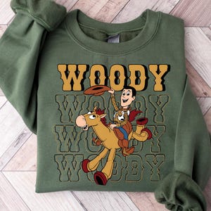May include: An olive green sweatshirt featuring a graphic of Woody from Toy Story riding a horse. The word "WOODY" is printed in a retro font above the graphic, repeated below. The sweatshirt has long sleeves and a crew neck.