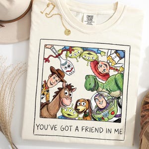 Puede incluir: Camiseta color crema con una ilustraci&oacute;n colorida de personajes de Toy Story, enmarcada en un borde negro. El texto "YOU'VE GOT A FRIEND IN ME" est&aacute; impreso debajo. La imagen se complementa con sandalias y un sombrero.