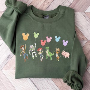 Puede incluir: Una sudadera de cuello redondo color verde oliva con un estampado gr&aacute;fico de seis personajes animados de Toy Story. Cada personaje sostiene una cuerda atada a un globo de colores con forma de Mickey Mouse. Los personajes incluyen a Woody, Buzz Lightyear, Jessie, Rex, Slinky Dog y Hamm.
