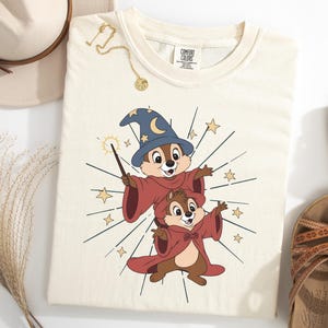 May include: Cream-colored t-shirt featuring a cartoon wizard design with two chipmunks wearing wizard robes and hats. The design includes stars and a magic wand. The shirt is styled with a hat, sandals, and jewelry.