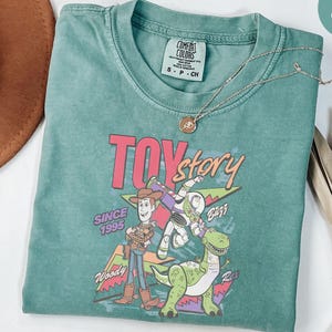 K&ouml;nnte beinhalten: Hellgr&uuml;nes T-Shirt mit einem Grafikdesign aus dem Film "Toy Story". Das Design zeigt Woody, Buzz Lightyear und Rex, mit dem Text "TOY STORY" und "Since 1995". Eine silberne Halskette mit Anh&auml;nger liegt auf dem Shirt.