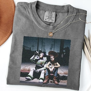 May include: A charcoal gray crew neck t-shirt featuring a graphic print of two animated characters sitting on a curb at night. The shirt has a Comfort Colors brand label at the inner neckline, indicating size S, and is made of 100% cotton fabric.