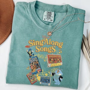 May include: A sage green short-sleeve t-shirt featuring a graphic print with the text Sing Along Songs, Disney characters, a cassette tape labeled Disneyland Fun, and musical notes. The shirt has a Comfort Colors brand tag at the inner neckline.