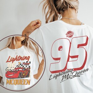 Op de afbeelding: Wit T-shirt met een Lightning McQueen-afbeelding. De voorkant toont "Lightning McQueen 95" met een rode raceauto en geblokte vlag. De achterkant heeft een grote rode "95" en "Lightning McQueen" tekst. Een vintage ontwerp.