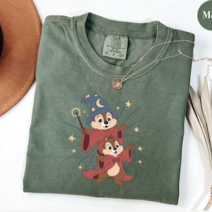 May include: Moss green t-shirt featuring a cartoon graphic of two chipmunks dressed as wizards, with a wand and stars. The shirt has a crew neck and short sleeves. A gold necklace with a pendant is draped across the shirt.