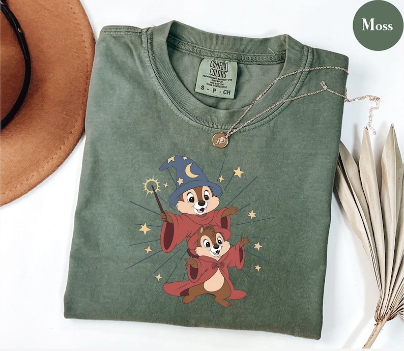 May include: Moss green t-shirt featuring a cartoon graphic of two chipmunks dressed as wizards, with a wand and stars. The shirt has a crew neck and short sleeves. A gold necklace with a pendant is draped across the shirt.