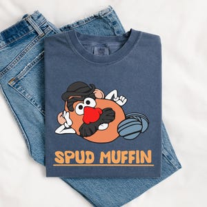 Puede incluir: Una camiseta azul marino con un personaje de dibujos animados de patata con sombrero negro, nariz roja y bigote. La camiseta tiene el texto "SPUD MUFFIN" en naranja. La camiseta est&aacute; doblada sobre un par de vaqueros azules.