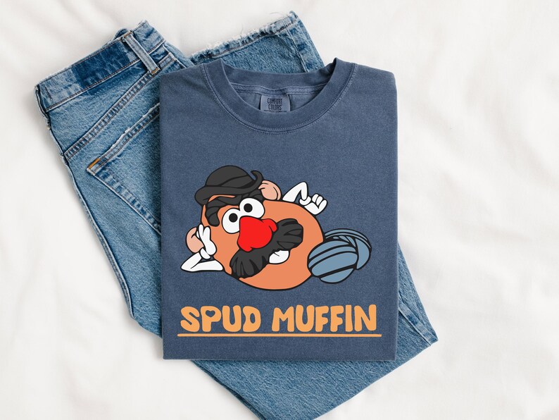 Puede incluir: Una camiseta azul marino con un personaje de dibujos animados de patata con sombrero negro, nariz roja y bigote. La camiseta tiene el texto "SPUD MUFFIN" en naranja. La camiseta est&aacute; doblada sobre un par de vaqueros azules.