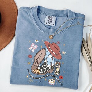 Puede incluir: Una camiseta azul de manga corta con un estampado gr&aacute;fico que muestra una bota de vaquero con estampado de vaca, un sombrero marr&oacute;n, bloques con las letras TS y el nombre Taylor. La camiseta tiene una etiqueta Comfort Colors y flores y estrellas.