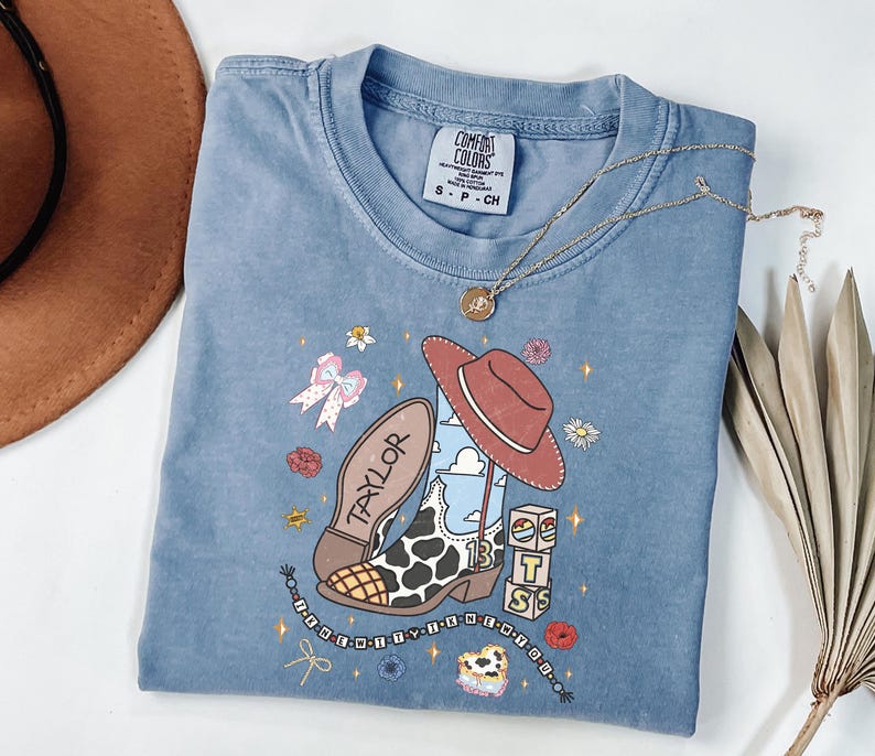 Puede incluir: Una camiseta azul de manga corta con un estampado gr&aacute;fico que muestra una bota de vaquero con estampado de vaca, un sombrero marr&oacute;n, bloques con las letras TS y el nombre Taylor. La camiseta tiene una etiqueta Comfort Colors y flores y estrellas.