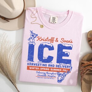 May include: A light pink t-shirt with the text "Kristoff & Sven's ICE" in blue and red. The shirt also features a graphic of Olaf the snowman and Sven the reindeer. The shirt is paired with brown sandals and a beige hat.