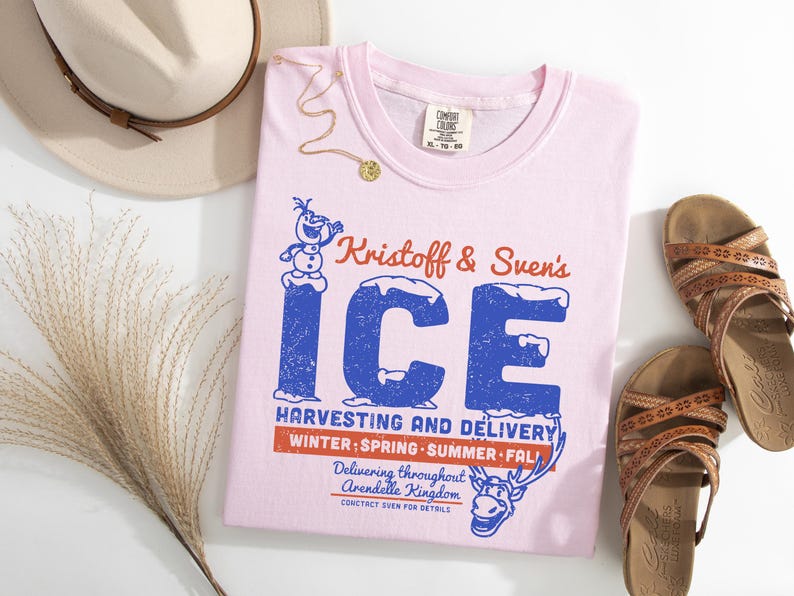 May include: A light pink t-shirt with the text "Kristoff & Sven's ICE" in blue and red. The shirt also features a graphic of Olaf the snowman and Sven the reindeer. The shirt is paired with brown sandals and a beige hat.