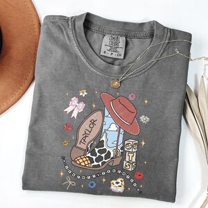 Puede incluir: Una camiseta de manga corta gris carb&oacute;n con un estampado gr&aacute;fico de una bota de vaquero con estampado de vaca, un sombrero rojo, bloques con las letras T, S y un arco&iacute;ris, y el texto Taylor e I KNEW YOU WERE TROUBLE, rodeado de iconos florales.