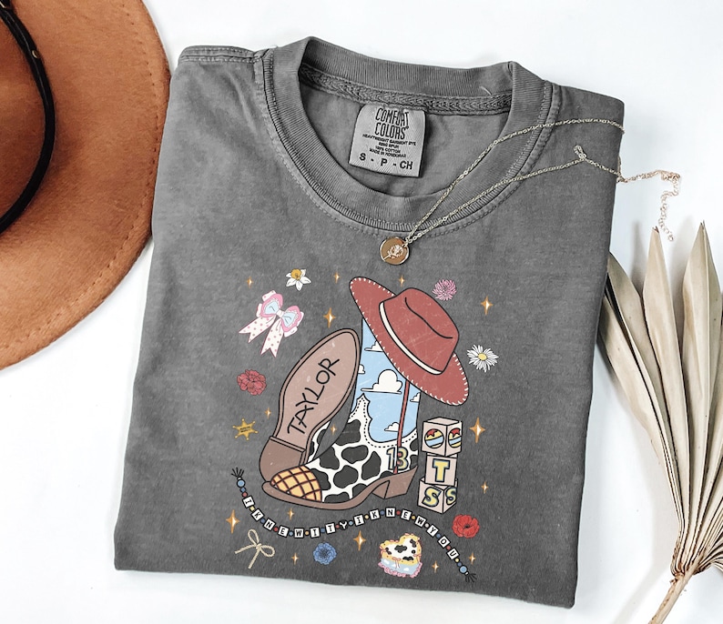 Puede incluir: Una camiseta de manga corta gris carb&oacute;n con un estampado gr&aacute;fico de una bota de vaquero con estampado de vaca, un sombrero rojo, bloques con las letras T, S y un arco&iacute;ris, y el texto Taylor e I KNEW YOU WERE TROUBLE, rodeado de iconos florales.