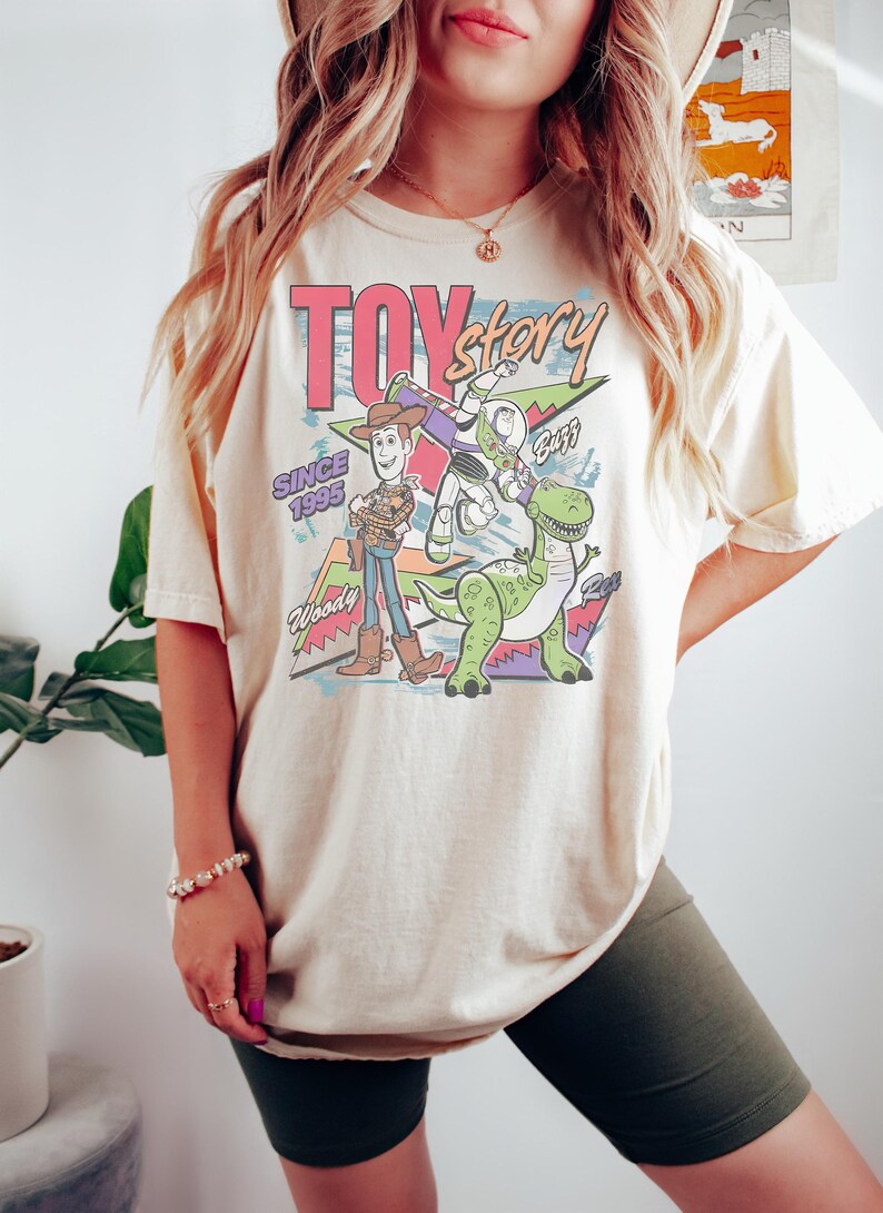 K&ouml;nnte beinhalten: Cremefarbenes T-Shirt mit einer farbenfrohen Grafik der Toy Story-Charaktere Woody, Buzz Lightyear und Rex. Der Text "Toy Story" ist &uuml;ber den Charakteren zu sehen, mit "Since 1995" und "Woody" ebenfalls sichtbar.