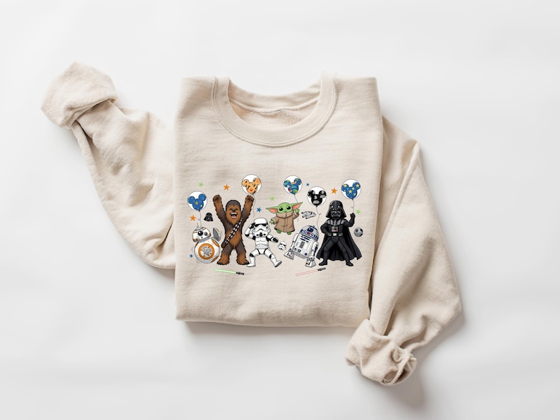 May include: A beige crewneck sweatshirt featuring a graphic print of Star Wars characters including Chewbacca, a Stormtrooper, Grogu, R2-D2, Darth Vader, and BB-8. The characters are holding balloons shaped like Mickey Mouse ears.