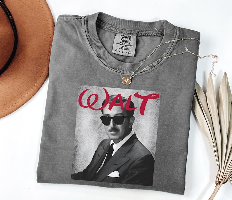Op de afbeelding: Grijs T-shirt met een zwart-wit afbeelding van Walt Disney met een zonnebril, met het woord "WALT" in rood script. Het shirt heeft een ronde hals en korte mouwen. Een gouden ketting is over het shirt gedrapeerd.