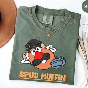Puede incluir: Una camiseta verde musgo con un personaje de dibujos animados de patata con sombrero negro y bigote, y una nariz roja. La camiseta tiene las palabras "SPUD MUFFIN" en naranja. Un collar dorado est&aacute; drapeado sobre la camiseta.