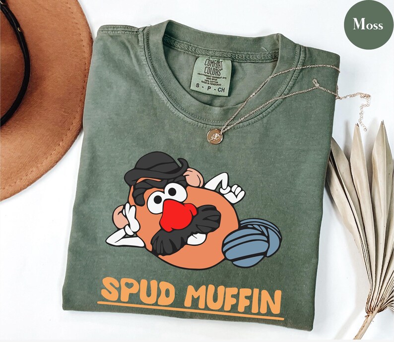 Puede incluir: Una camiseta verde musgo con un personaje de dibujos animados de patata con sombrero negro y bigote, y una nariz roja. La camiseta tiene las palabras "SPUD MUFFIN" en naranja. Un collar dorado est&aacute; drapeado sobre la camiseta.