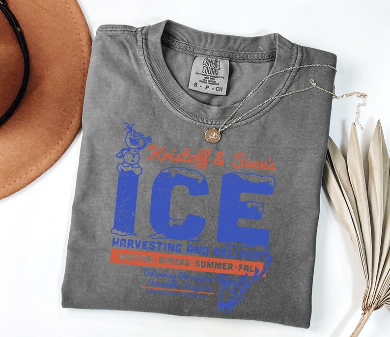 May include: A gray Comfort Colors t-shirt with a blue and orange graphic that reads "Kristoff & Sven's ICE Harvesting and..." The shirt has a crew neck and short sleeves. A silver necklace with a pendant is on the shirt.