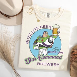 May include: Cream-colored t-shirt featuring a graphic of Buzz Lightyear holding a beer, with text reading "Buzz Lite Beer Star Command Brewery." The design includes a blue circle and the words "Best Quality Since 1995."