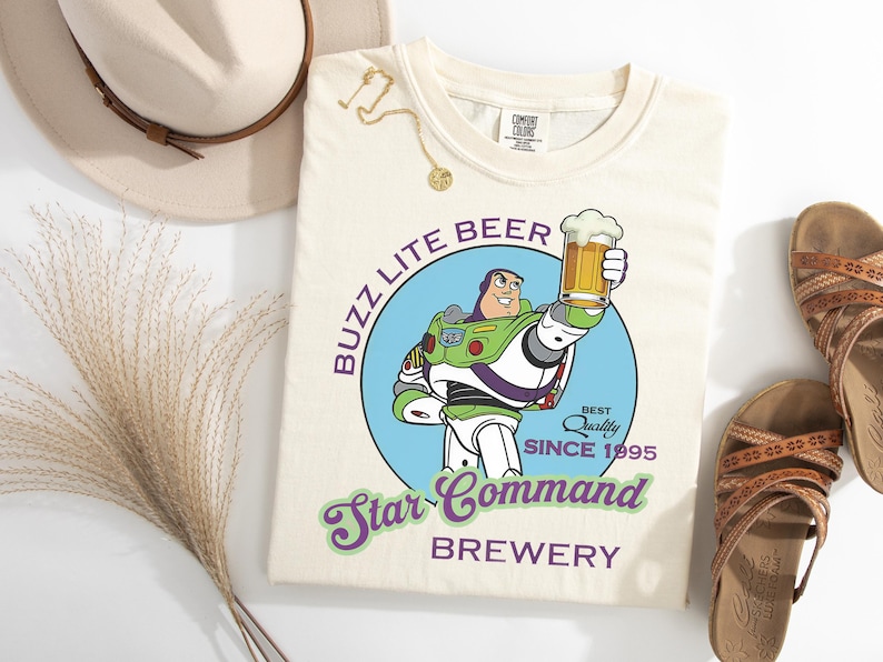 May include: Cream-colored t-shirt featuring a graphic of Buzz Lightyear holding a beer, with text reading "Buzz Lite Beer Star Command Brewery." The design includes a blue circle and the words "Best Quality Since 1995."