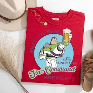 May include: A red t-shirt featuring a cartoon image of Buzz Lightyear holding a beer mug, with the text "Buzz Lite Beer Star Command Brewery Since 1995". The shirt is accompanied by a hat, sandals, and a necklace.