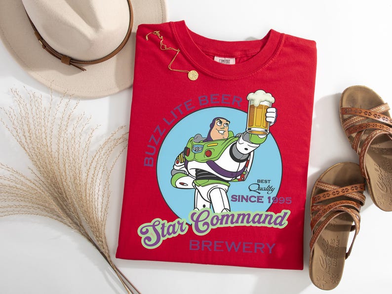 May include: A red t-shirt featuring a cartoon image of Buzz Lightyear holding a beer mug, with the text "Buzz Lite Beer Star Command Brewery Since 1995". The shirt is accompanied by a hat, sandals, and a necklace.