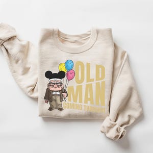 Op de afbeelding: Beige sweatshirt met de gouden tekst "OLD MAN COMING THROUGH". Het heeft een cartoon illustratie van een oude man met ballonnen. De man draagt een bril, een bruine jas en een zwarte Mickey Mouse hoed.