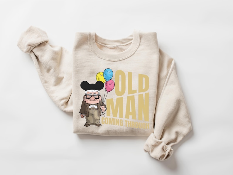 Op de afbeelding: Beige sweatshirt met de gouden tekst "OLD MAN COMING THROUGH". Het heeft een cartoon illustratie van een oude man met ballonnen. De man draagt een bril, een bruine jas en een zwarte Mickey Mouse hoed.