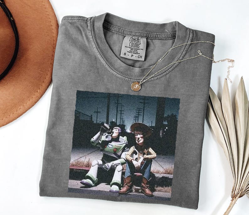 May include: A charcoal gray crew neck t-shirt featuring a graphic print of two animated characters sitting on a curb at night. The shirt has a Comfort Colors brand label at the inner neckline, indicating size S, and is made of 100% cotton fabric.