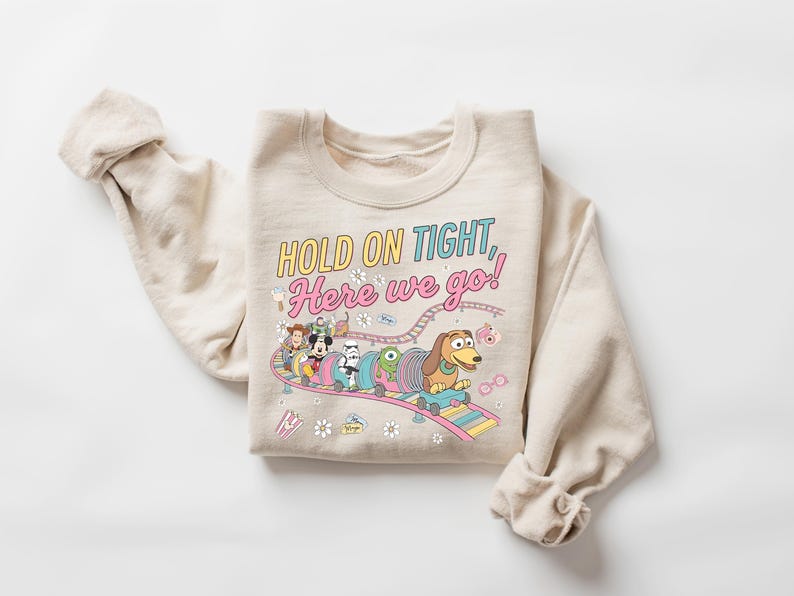 May include: A beige crewneck sweatshirt featuring a colorful graphic of various cartoon characters riding a roller coaster. The text reads, "HOLD ON TIGHT, Here we go!" The design includes small illustrations of flowers, popcorn, and glasses surrounding the coaster track.