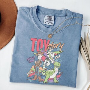 K&ouml;nnte beinhalten: Ein blaues kurz&auml;rmeliges T-Shirt mit dem Toy Story-Logo und den Figuren Woody, Buzz Lightyear und Rex. Das Shirt hat einen Rundhalsausschnitt und ein Comfort Colors-Etikett. Eine goldene Halskette mit Anh&auml;nger liegt &uuml;ber dem Shirt.
