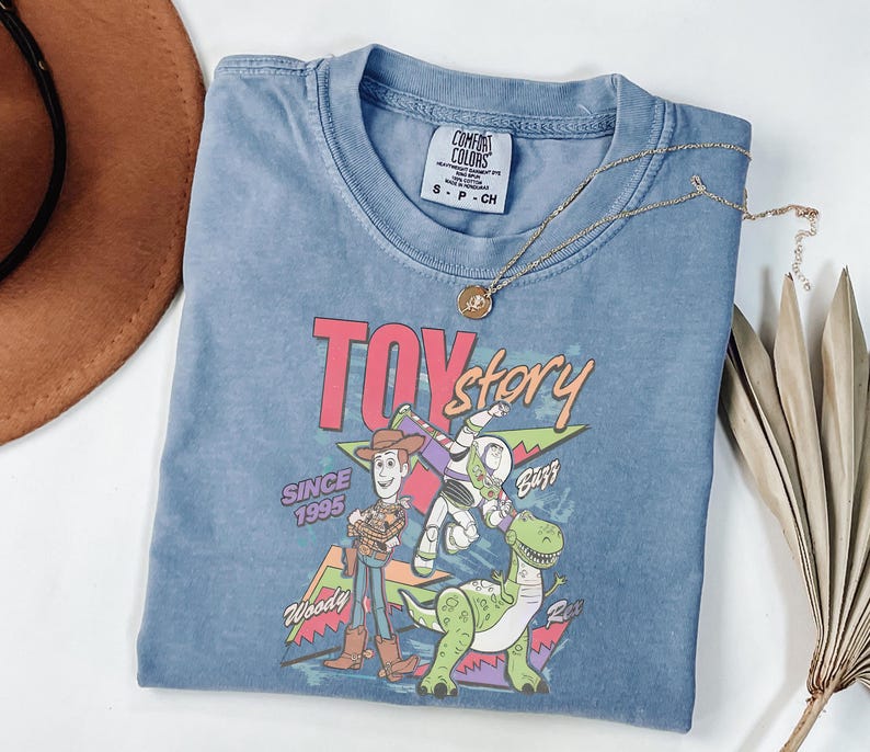 K&ouml;nnte beinhalten: Ein blaues kurz&auml;rmeliges T-Shirt mit dem Toy Story-Logo und den Figuren Woody, Buzz Lightyear und Rex. Das Shirt hat einen Rundhalsausschnitt und ein Comfort Colors-Etikett. Eine goldene Halskette mit Anh&auml;nger liegt &uuml;ber dem Shirt.
