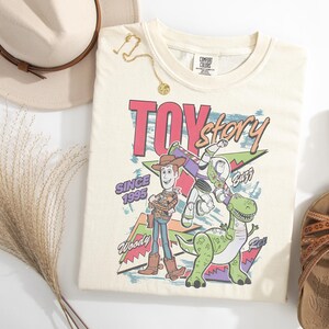 K&ouml;nnte beinhalten: Cremefarbenes T-Shirt mit dem Schriftzug "Toy Story" in Retro-Schrift, mit Woody, Buzz Lightyear und Rex dem Dinosaurier. Das Shirt tr&auml;gt auch den Text "Since 1995". Accessoires sind ein Hut, Sandalen und eine goldene Halskette.