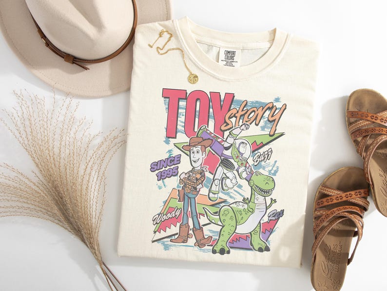 K&ouml;nnte beinhalten: Cremefarbenes T-Shirt mit dem Schriftzug "Toy Story" in Retro-Schrift, mit Woody, Buzz Lightyear und Rex dem Dinosaurier. Das Shirt tr&auml;gt auch den Text "Since 1995". Accessoires sind ein Hut, Sandalen und eine goldene Halskette.
