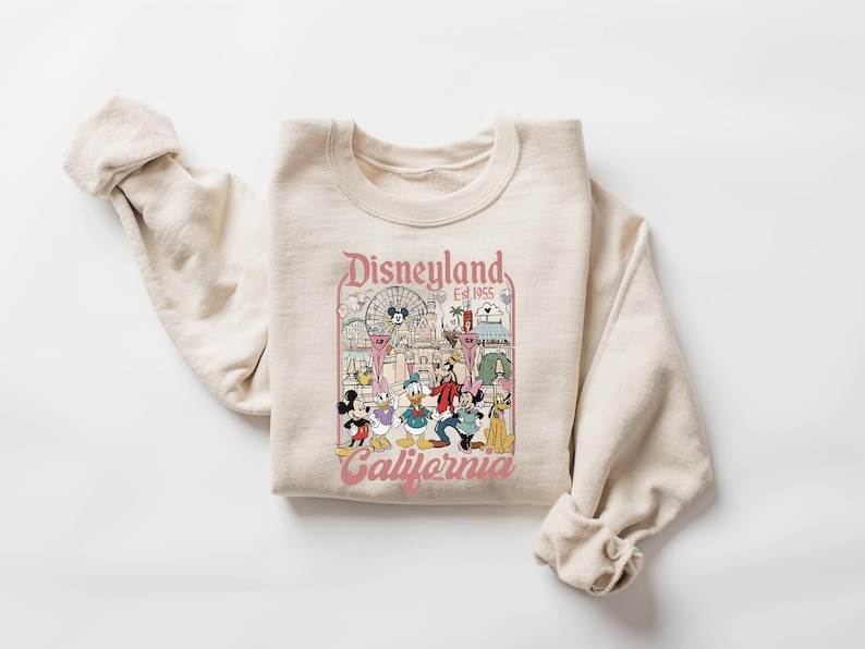 May include: A beige crewneck sweatshirt featuring a graphic print with the text Disneyland Est. 1955 California. The design includes illustrations of Mickey Mouse, Minnie Mouse, Donald Duck, Goofy, and Pluto in front of a park castle and ferris wheel.
