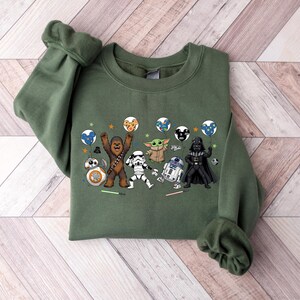 May include: An olive green crewneck sweatshirt featuring a graphic print of Star Wars characters including Chewbacca, a Stormtrooper, Grogu, R2-D2, Darth Vader, and BB-8. The characters are holding colorful balloons and are surrounded by small stars.