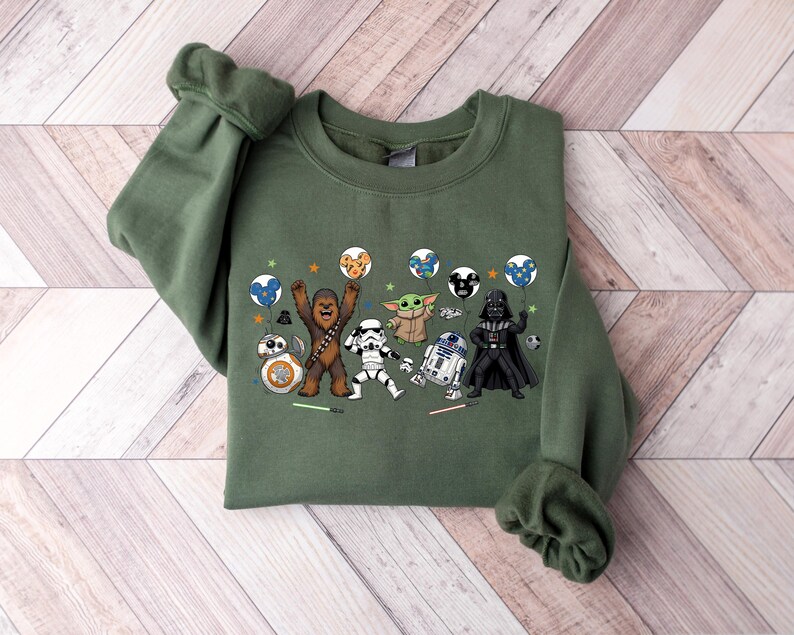 May include: An olive green crewneck sweatshirt featuring a graphic print of Star Wars characters including Chewbacca, a Stormtrooper, Grogu, R2-D2, Darth Vader, and BB-8. The characters are holding colorful balloons and are surrounded by small stars.